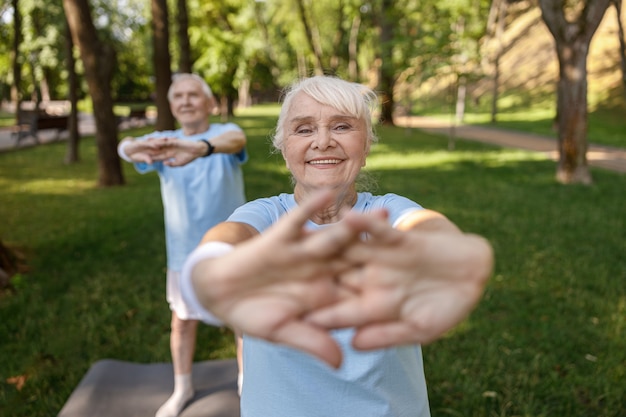 Joyful senior lady does sports exercise training with friend on lawn in city park