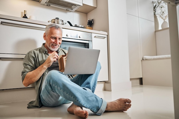 Photo joyful senior man with gift and laptop sitting on floor