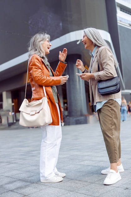 Joyful senior women with mobile phones laugh standing on city street on autumn day