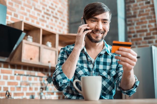 Joyful smart positive man talking on the phone and smiling while looking at his credit card