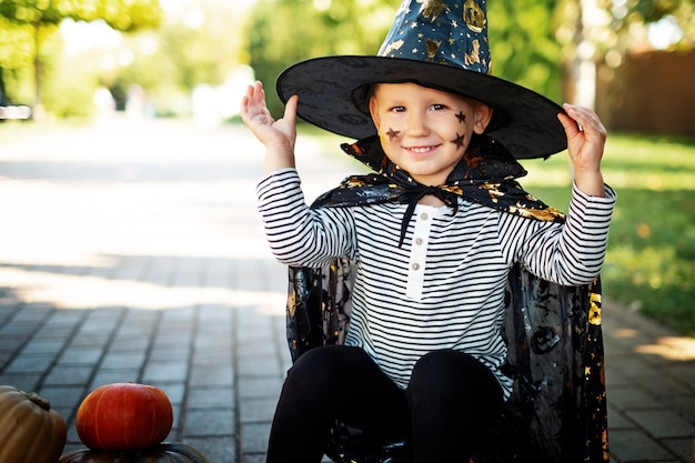 joyful smiling child dressed as wizard in hat and raincoat sits on poke on the street