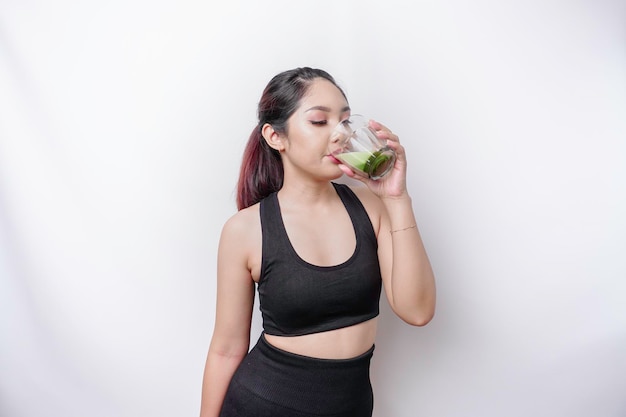 Joyful sporty Asian woman wearing sportswear is drinking a glass of tasty green smoothie isolated on white background