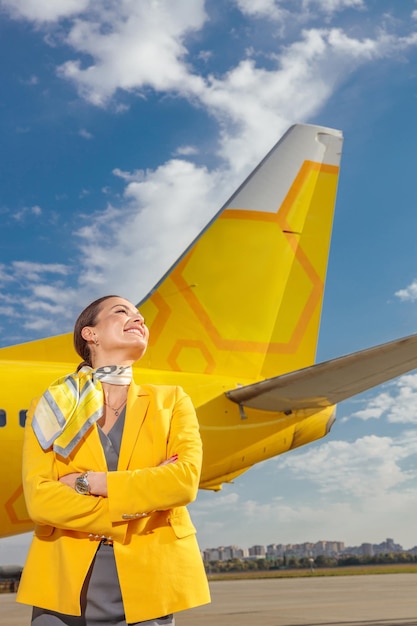 Joyful stewardess in airline uniform keeping arms crossed and smiling while standing near airplane at airport