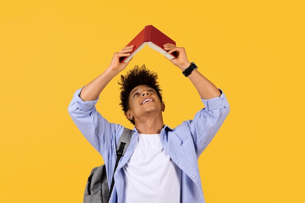 Joyful student with book roof over head on yellow background