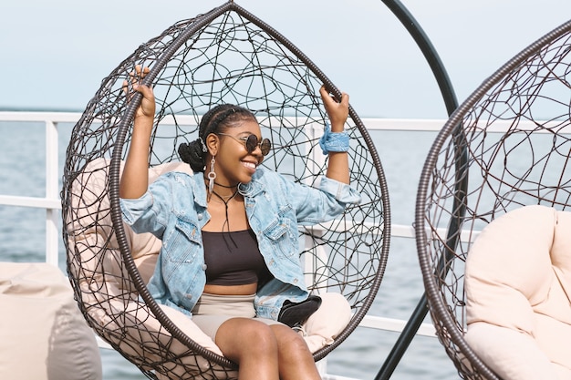 Joyful stylish young woman in denim jacket, sunglasses and long earrings sitting in hanging chair at outdoor birthday party