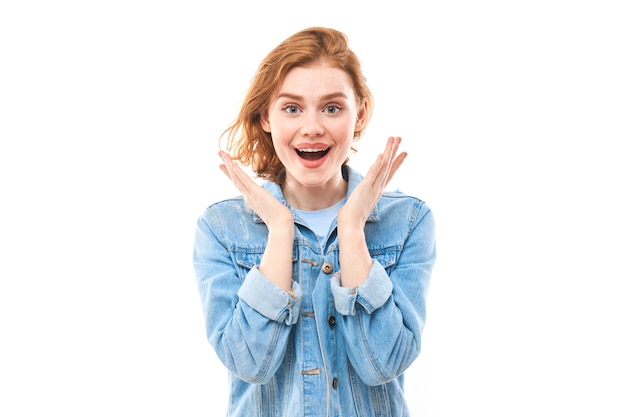 Joyful surprised redhead girl looks at the camera. Excited young woman on a white background in a blue denim jacket