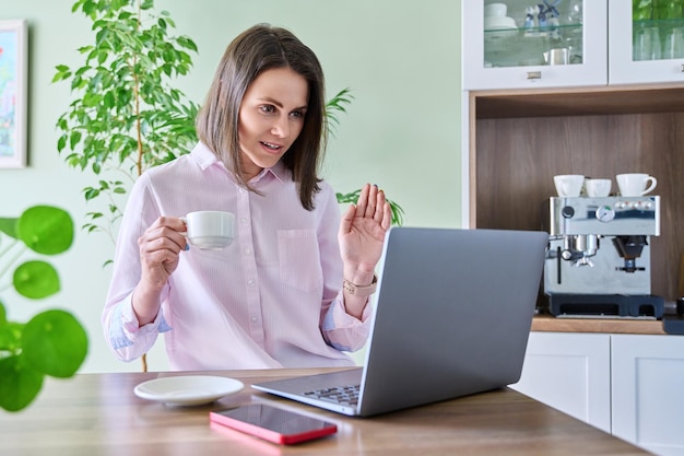 Joyful surprised woman looking at laptop screen sitting at home in kitchen