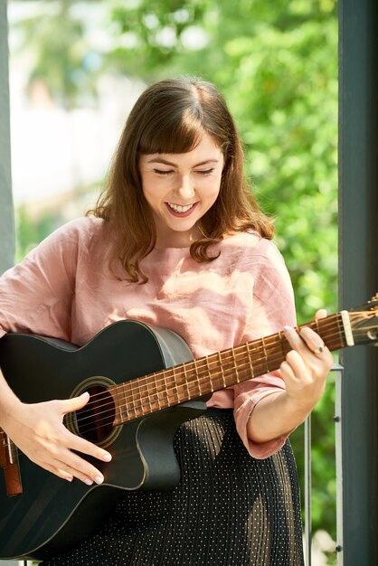 Joyful talented young woman enjoying playing guitar and singing when standing on house porch