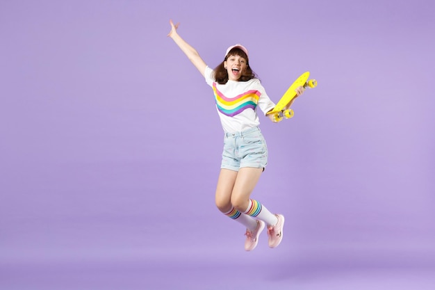 Радостная девочка-подросток в яркой одежде держит желтый скейтборд, веселится, прыгает, расправляет руки, изолированные на фиолетовом пастельном фоне. Люди искренние эмоции, концепция образа жизни. Скопируйте пространство для копирования.