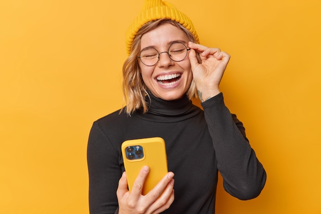 Photo joyful teenage girl watches funny video content via cellphone laughs happily keeps hand on rim of spectacles wears hat and black poloneck poses against yellow background checks positive email