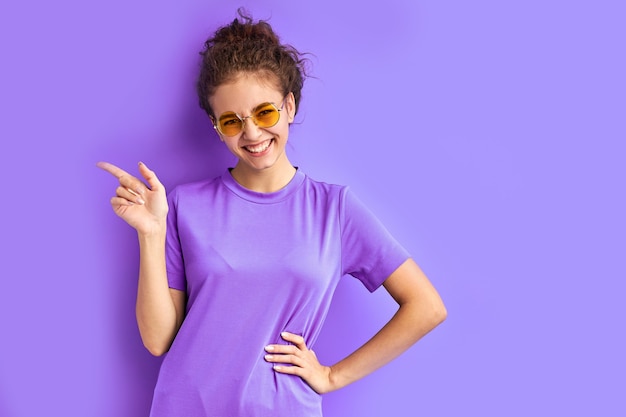 Joyful teenager girl in t-shirt and sunglasses posing at camera, pointing fingers at side, index at