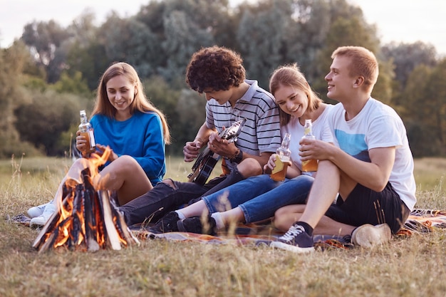 Joyful teenagers sit closely to each other on ground near fire, have picnic together, play acoustic guitar, celebrate something