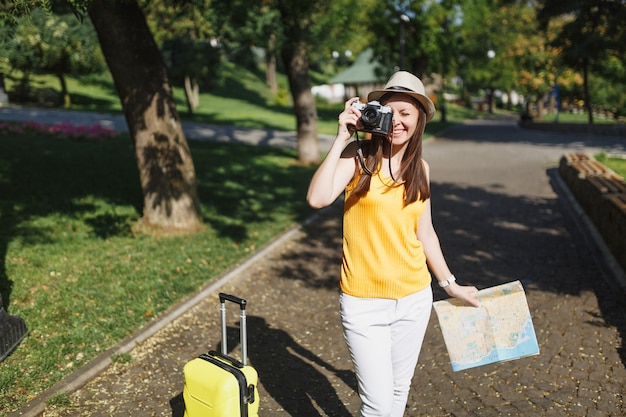 スーツケースの都市地図と帽子をかぶったうれしそうな旅行者の観光客の女性は、都市の屋外でレトロなビンテージ写真カメラで写真を撮ります。週末の休暇で旅行するために海外旅行する女の子。観光の旅のライフスタイル。