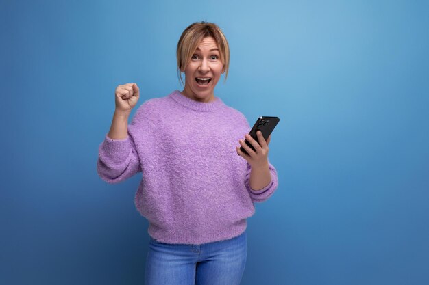 Photo joyful winning blond woman in purple hoodie having fun holding smartphone in hands on blue