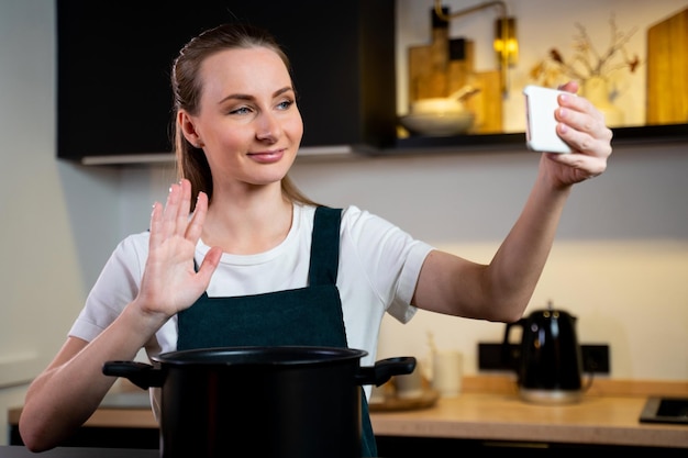Радостная женщина в фартуке готовит дома и пробует блюдо, снимая видеоурок по телефону в т