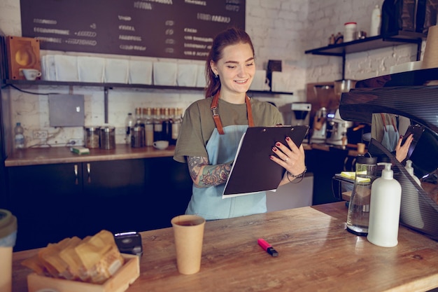 Joyful woman barista writing on clipboard in coffee shop