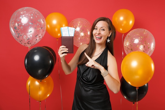 Joyful woman in black dress celebrating, pointing index finger on passport, boarding pass tickets in hand on bright red background air balloons. Happy New Year, birthday mockup holiday party concept.