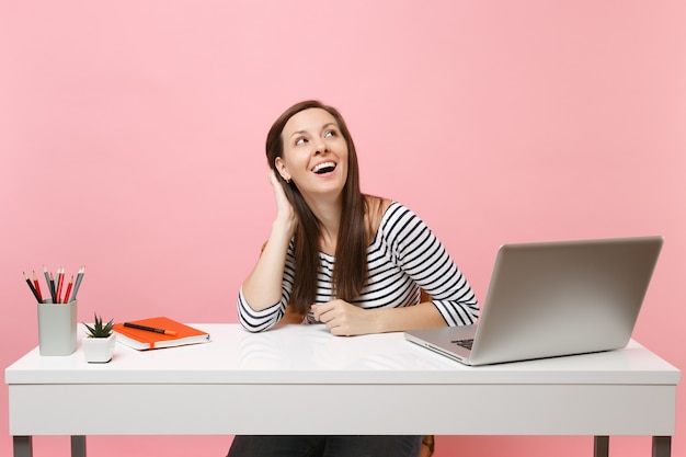 Donna gioiosa che tiene la mano sulla testa alzando lo sguardo pensando di sognare seduta a lavorare alla scrivania bianca con un computer portatile contemporaneo