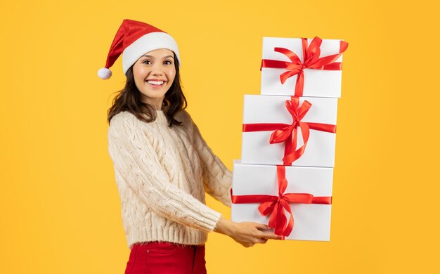 Joyful woman in santa hat holding xmas gifts yellow backdrop