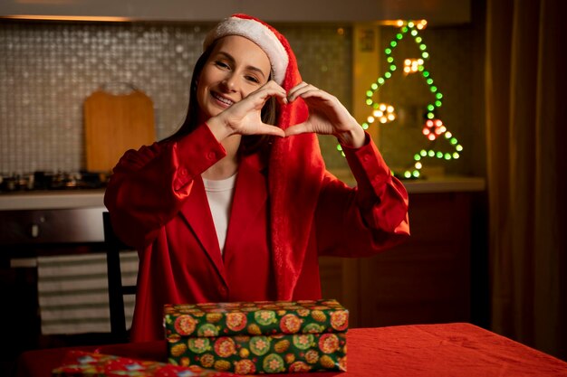 A joyful woman wearing a santa hat creates a heart sign with her hands while standing behind a