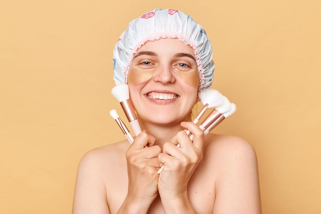 Joyful woman wearing shower cap standing isolated over beige background posing with big variety of makeup brushes doing beauty routine procedures after taking shower