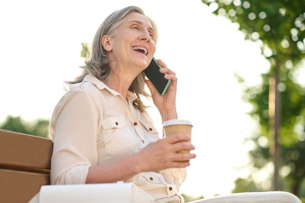 Joyful woman with coffee talking on smartphone