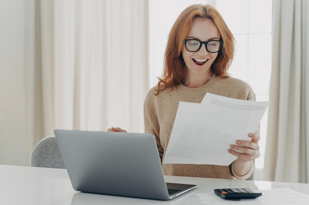 Joyful woman with glasses holding papers celebrating mortgage closure near laptop