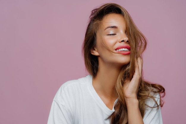 Joyful woman with hand on hair white tee closed eyes pink background