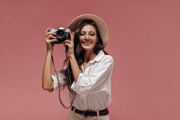 Joyful woman with long black hair in clear eyeglasses white shirt and light boater looking into camera and makes photo