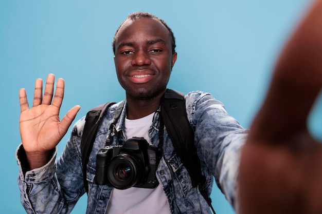 Joyful young adult waving at camera while having professional camera and holiday trip backpack on blue background. Photography enthusiast with DSLR photo device taking picture of himself.