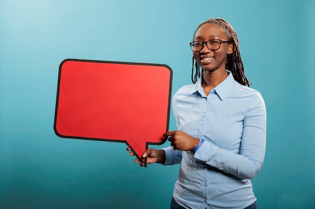 Joyful young adult woman holding blank template announcement placard. African american confident and smiling heartily woman holding red cardboard speech bubble on blue background.