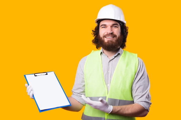 A joyful young architect is looking and smiling at the camera and holding his work pappers is pointing at them near a yellow wall