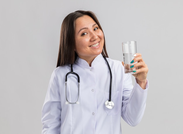 Joyful young asian female doctor wearing medical robe and stethoscope holding glass of water  isolated on white wall