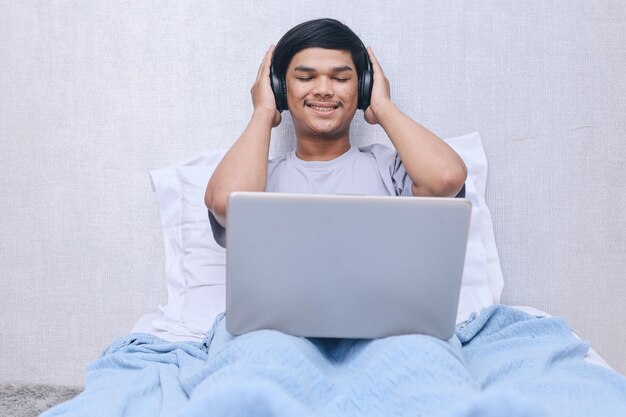 Joyful young Asian man wearing headphones and listening music from laptop while sitting on bed at be