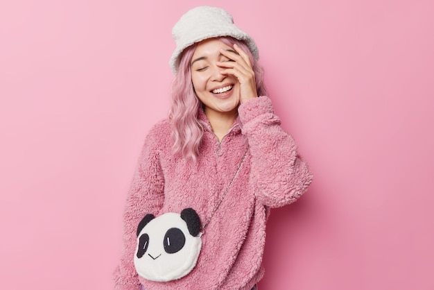 Joyful young Asian woman covers half of face with hand smiles happily keeps eyes closed smiles pleasantly feels pleased wears winter hat and coat isolated over pink background. Emotions concept