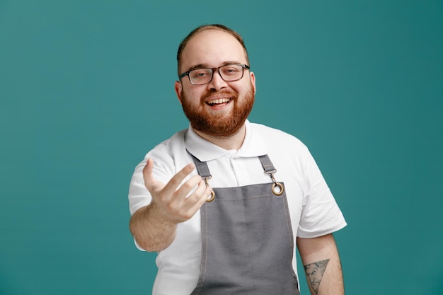 Joyful young barber wearing uniform and glasses looking at camera showing come here gesture isolated on blue background