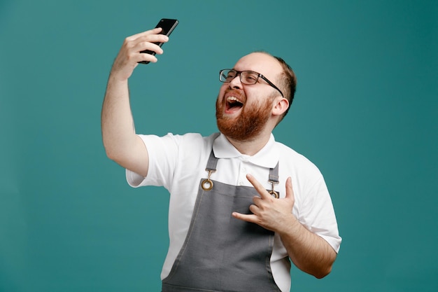 Joyful young barber wearing uniform and glasses raising mobile phone up showing rock sign taking selfie isolated on blue background