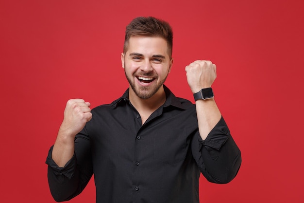 Joyful young bearded guy 20s in classic black shirt posing isolated on red background studio portrait. People lifestyle concept. Mock up copy space. Wearing smart watch on hand, doing winner gesture.