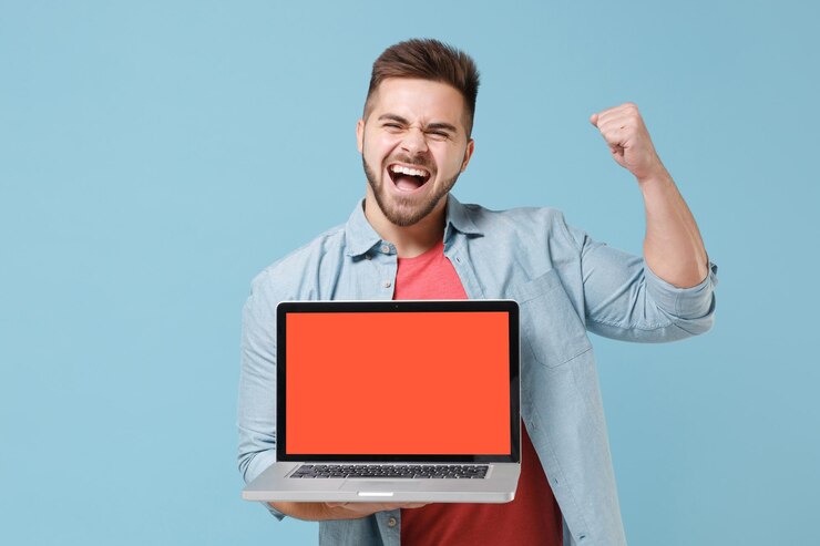 फोटो joyful young bearded guy in casual shirt posing isolated on pastel blue background. people lifestyle concept. mock up copy space. hold laptop pc computer with blank empty screen doing wwinner gesture.