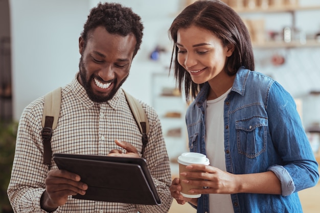 Joyful young best friends standing in the coffeehouse and checking its menu on the tablet while the woman holding a cup of coffee