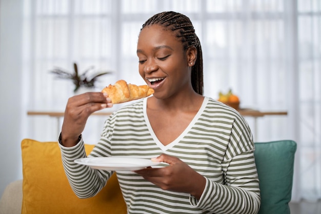 Joyful young black lady having breakfast at home eating croissant