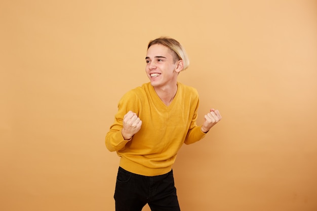 Радостный молодой блондин, одетый в желтый свитер, позирует в студии на бежевом фоне.