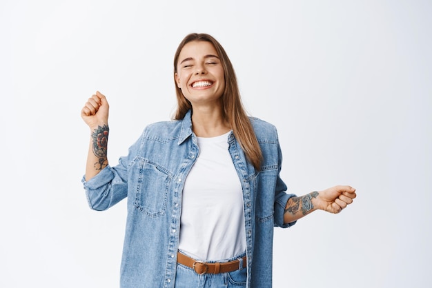 Joyful young blond woman having fun dancing with happy smiling face and relaxed emotion standing in casual denim shirt and white tshirt against white background