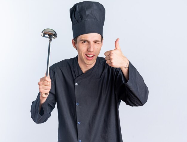 Joyful young blonde male cook in chef uniform and cap holding ladle winking showing thumb up 