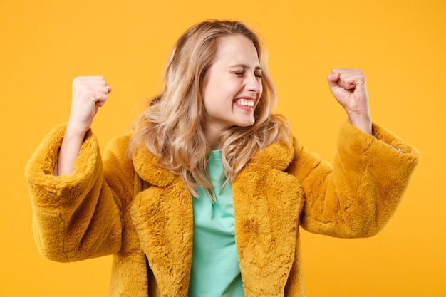 Joyful young blonde woman girl in yellow fur coat posing isolated on orange background studio portrait. People lifestyle concept. Mock up copy space. Keeping eyes closed, clenching fists like winner.