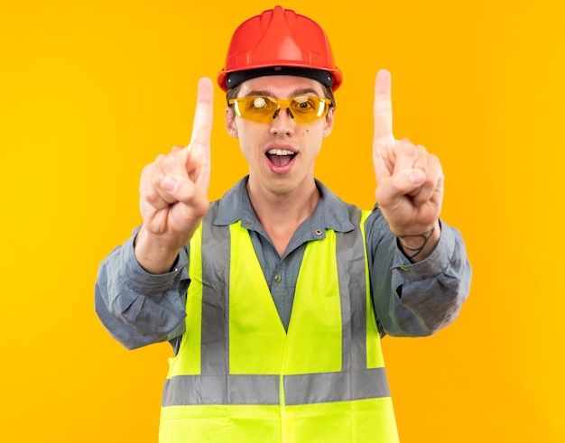 Joyful young builder man in uniform wearing glasses showing one 