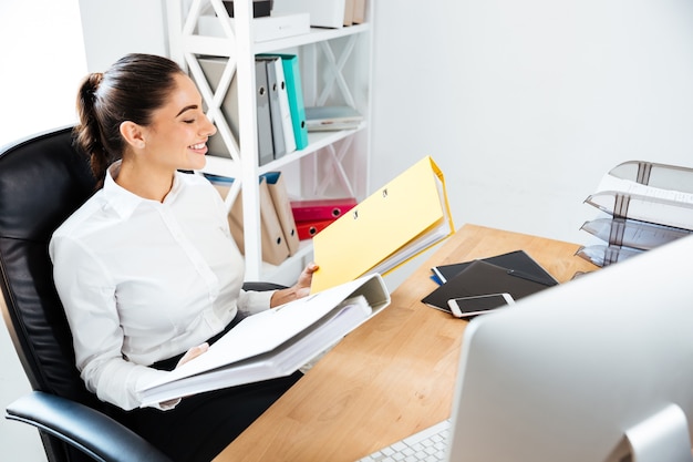 Joyful young businesswoman looking at yellow folders while sitting at the office desk