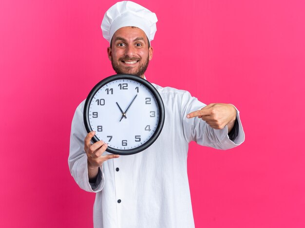 Joyful young caucasian male cook in chef uniform and cap showing clock pointing at it 