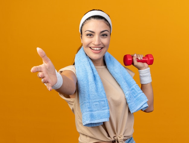 Joyful young caucasian sporty girl wearing headband and wristbands raising dumbbell looking at front stretching out hand towards camera with towel around neck isolated on orange wall