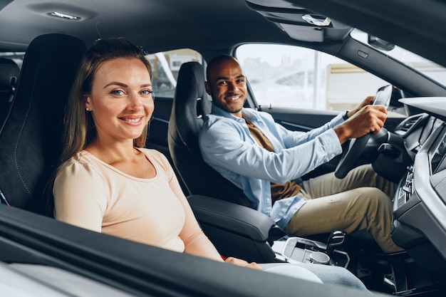 Gioiosa coppia giovane guardandosi intorno all'interno di una nuova auto che stanno per acquistare in una concessionaria di un negozio di auto